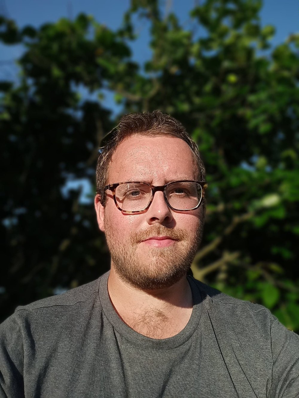 Tristan Hotham in tortoiseshell glasses and a grey t-shirt outdoors with green trees behind