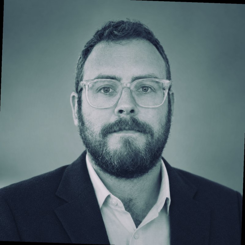 Black and white headshot of Tristan Hotham in clear glasses and a navy blazer