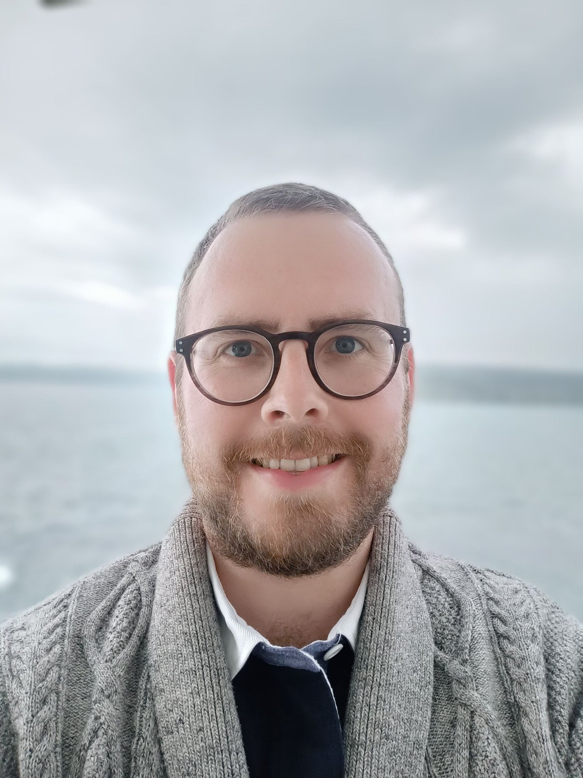 Portrait of Dr Tristan Hotham, smiling, wearing round tortoiseshell glasses and a grey cable-knit cardigan, with the sea behind