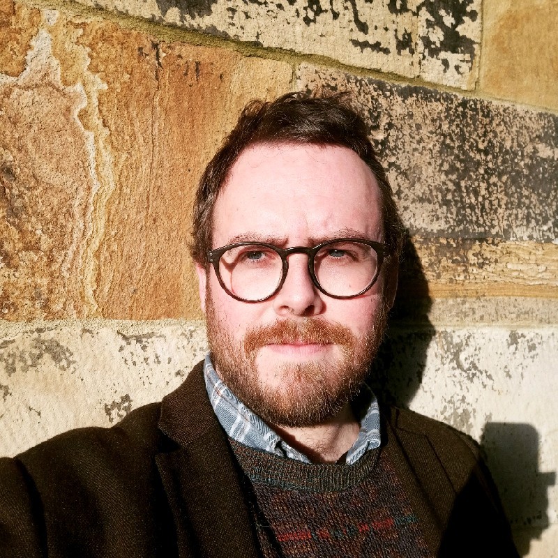 Tristan Hotham in round tortoiseshell glasses, a brown jacket, checked shirt and patterned jumper, against a sunlit sandstone wall