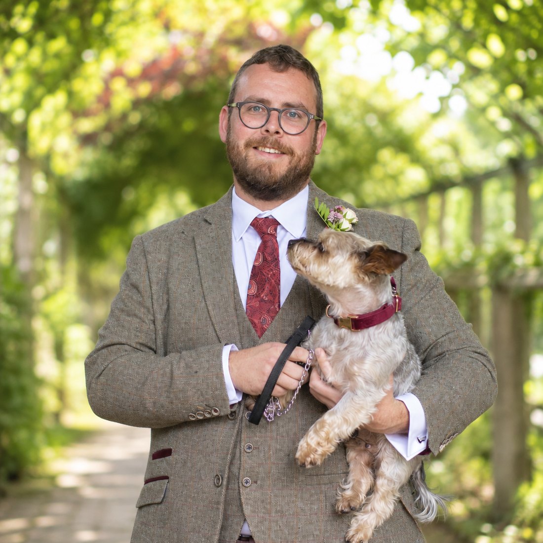 Tristan Hotham in a tweed suit and burgundy tie holding his terrier dog in a garden